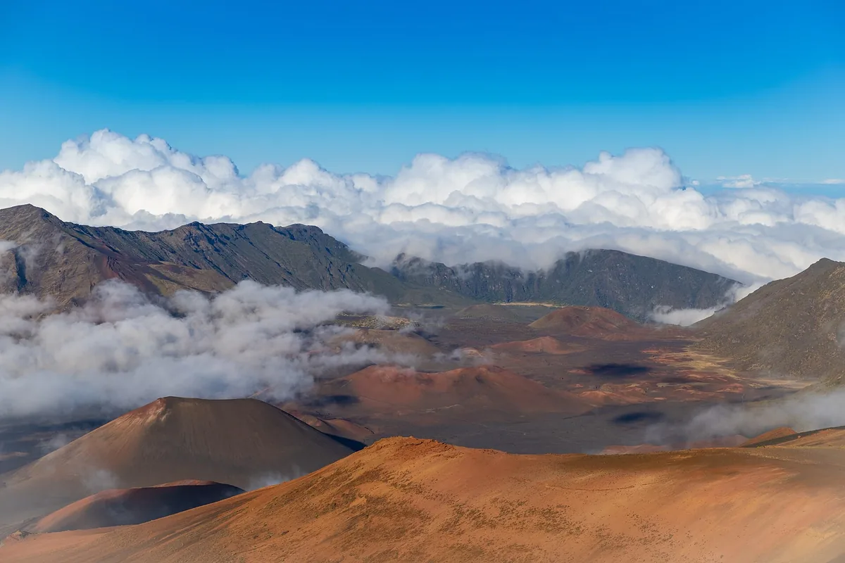 Red and brown cinder cones on the floor of Haleakala crater on Maui, with clouds spilling over the rim