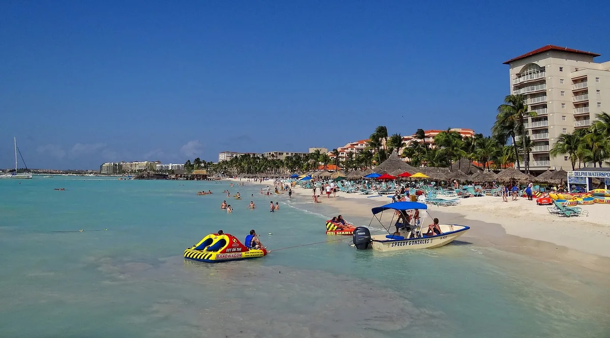 Swimmers, boats and a row of palm-shaded beach umbrellas along Palm Beach on Aruba, backed by high-rise hotels