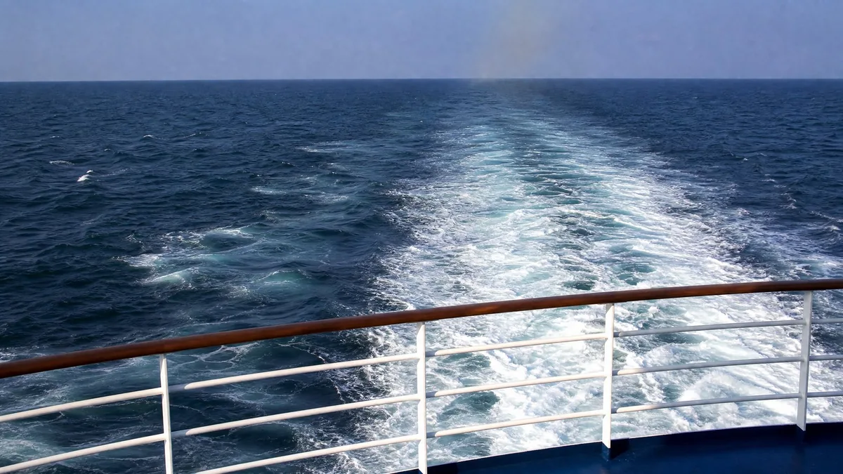 The white wake of a ferry on a deep blue sea seen from the rear deck railing, distant hazy horizon only