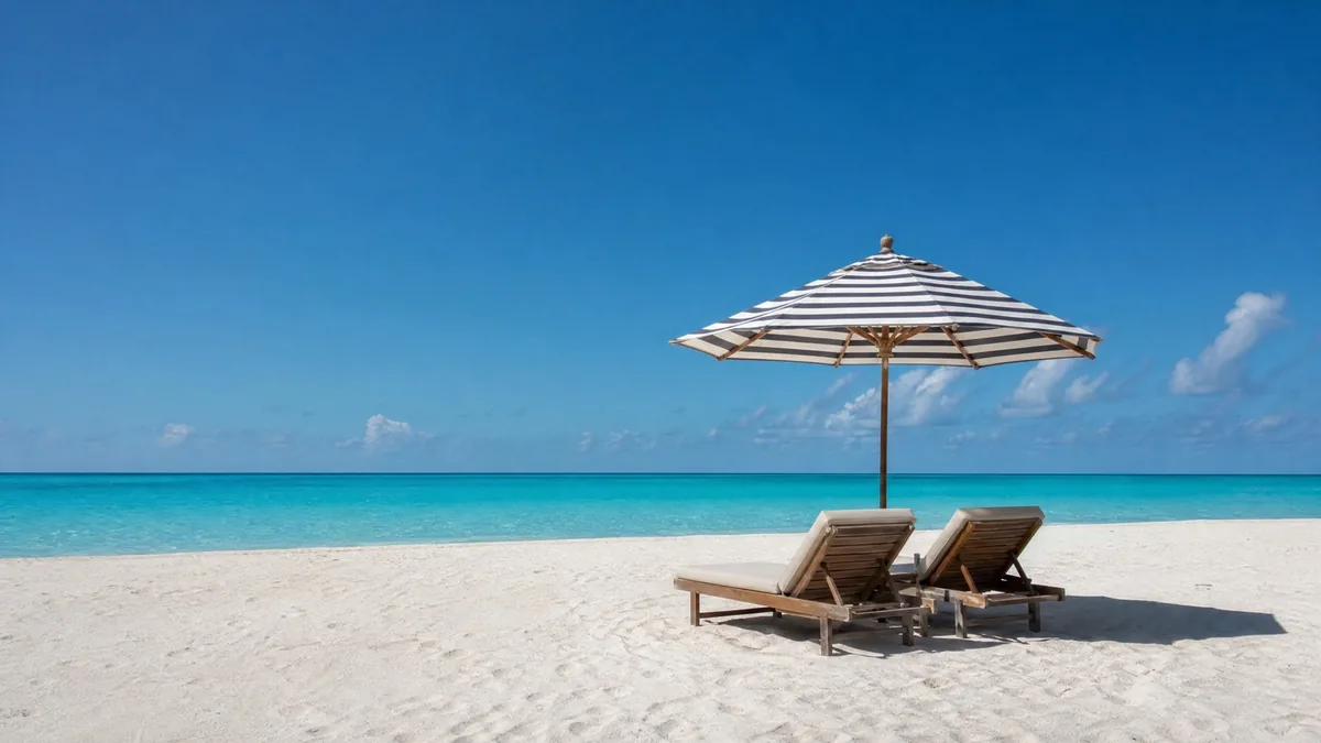 A single striped beach umbrella and two empty lounge chairs on a wide white beach, turquoise water, clear sky