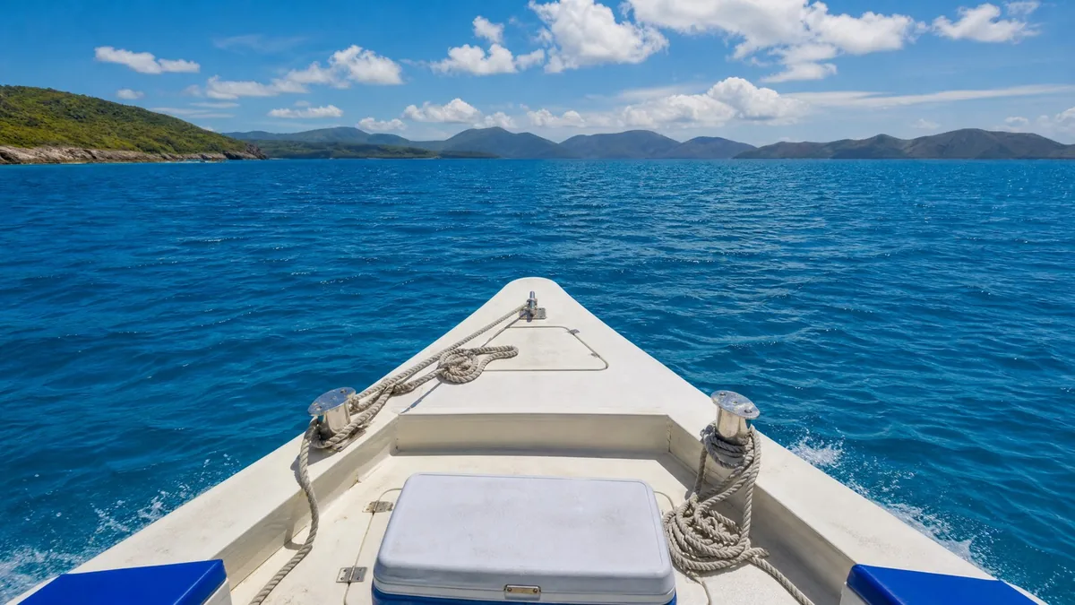 The bow of a small tour boat cutting through bright blue water on a sunny day, coiled ropes and a cooler on the deck