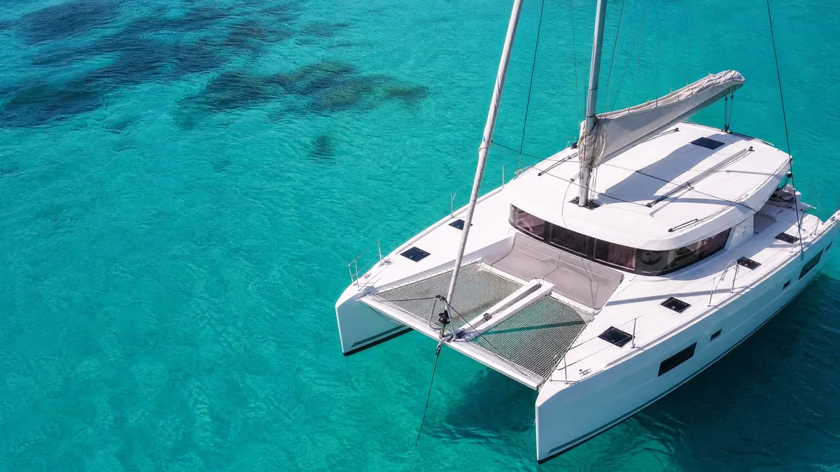 A white sailing catamaran anchored in calm turquoise water, its trampoline net and furled sail in view, seen from a little above