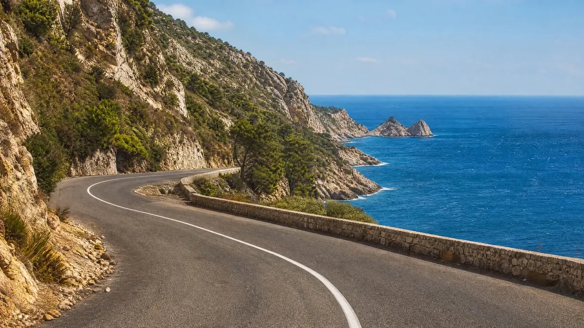 A winding two-lane coastal road with a low stone wall along cliffs above a blue sea