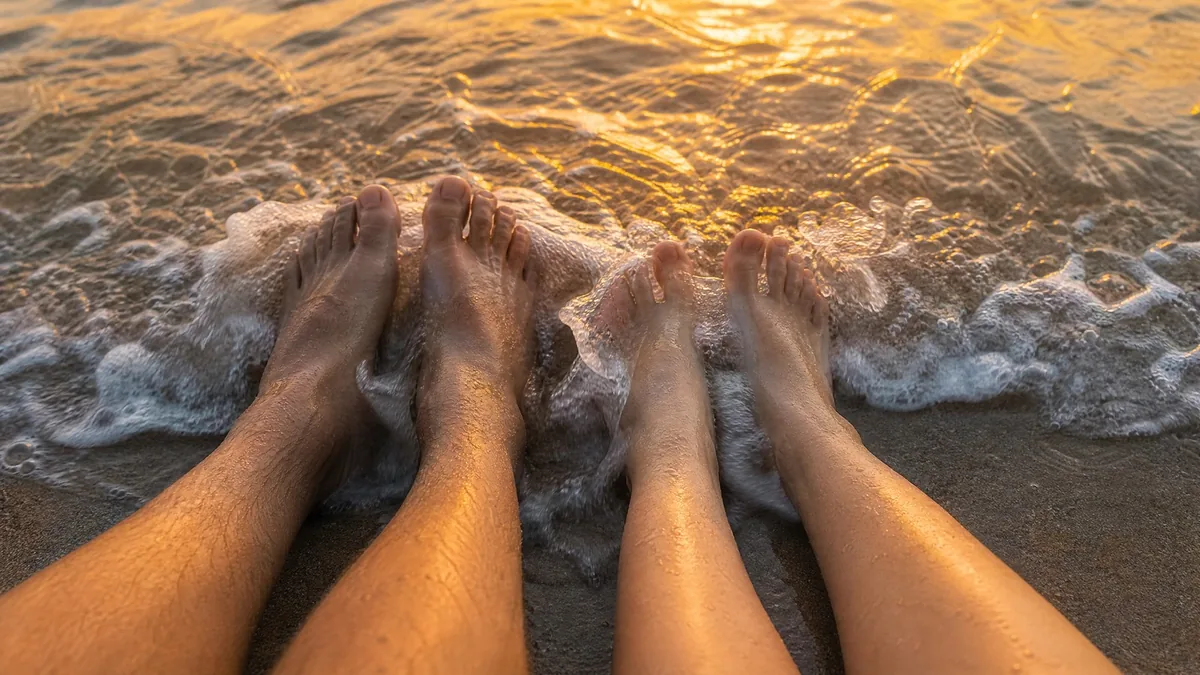 Two pairs of bare feet side by side on wet sand at sunset, a wave washing over them