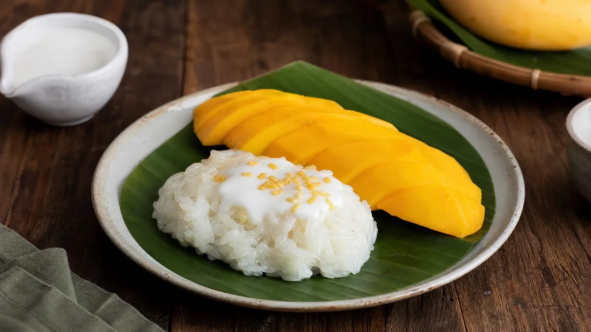 A plate of mango sticky rice with coconut cream on a banana leaf, on a wooden table