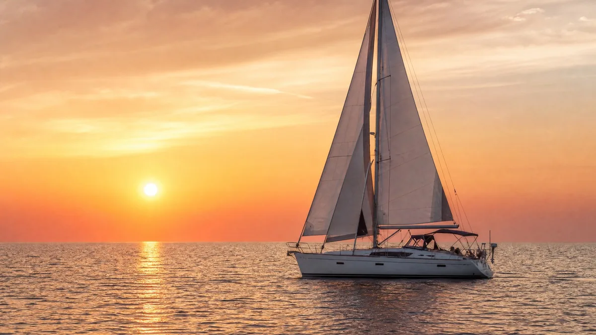 A white sailboat on a calm sea at sunset, orange and pink sky, golden water