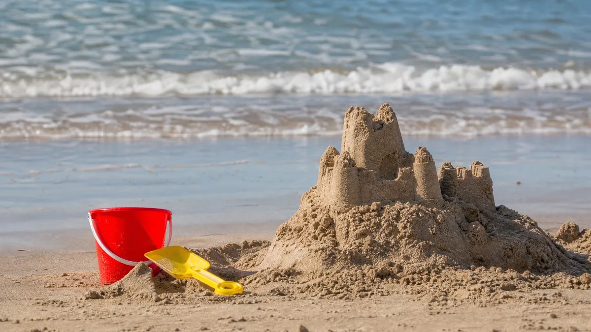 A child's sandcastle with a red plastic bucket and yellow spade on a beach, gentle waves behind
