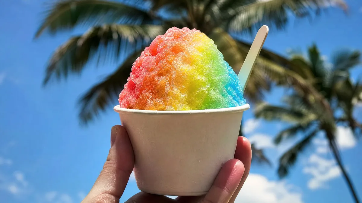A rainbow shave ice in a paper cup with a small spoon, held up against a blurred blue sky and palms, hand partly visible