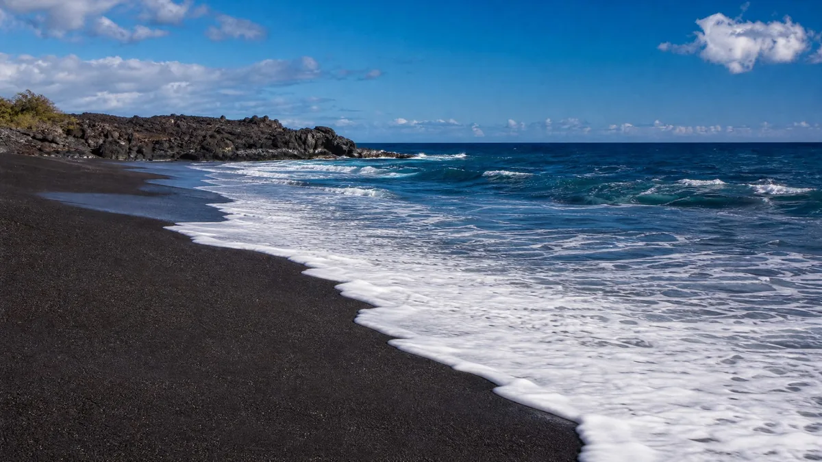 A black volcanic-sand beach with bright white surf and a deep blue sea