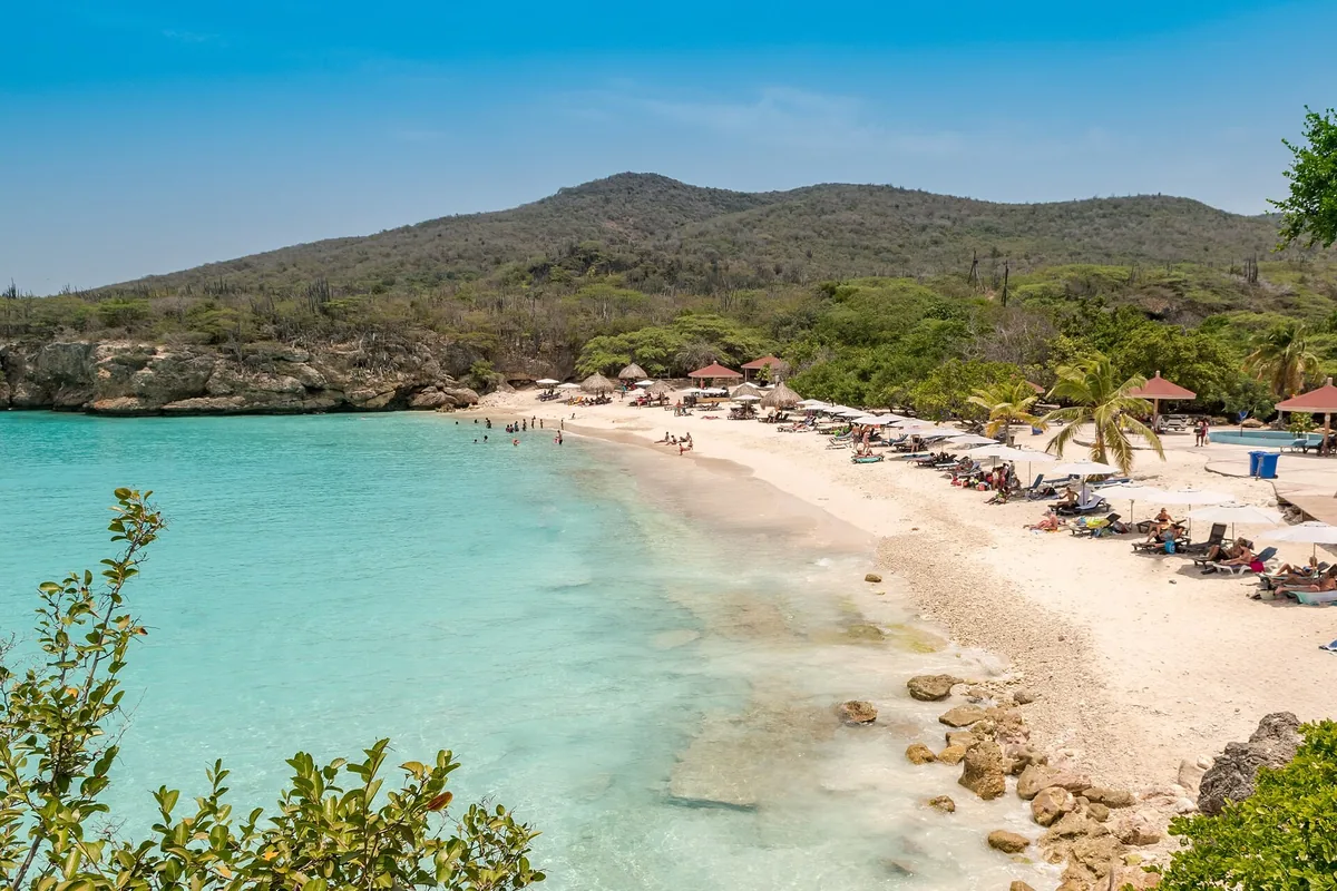 Turquoise water and white sand lined with umbrellas at Grote Knip (Playa Kenepa Grandi) beneath green hills on Curaçao
