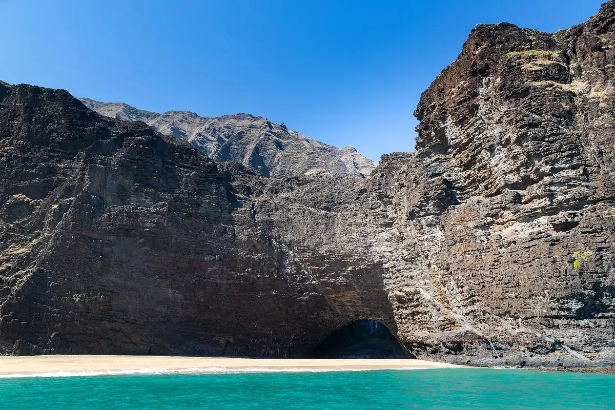 Towering dark sea cliffs and the Honopu natural arch above a strip of sand and turquoise water on Kauai's Na Pali Coast