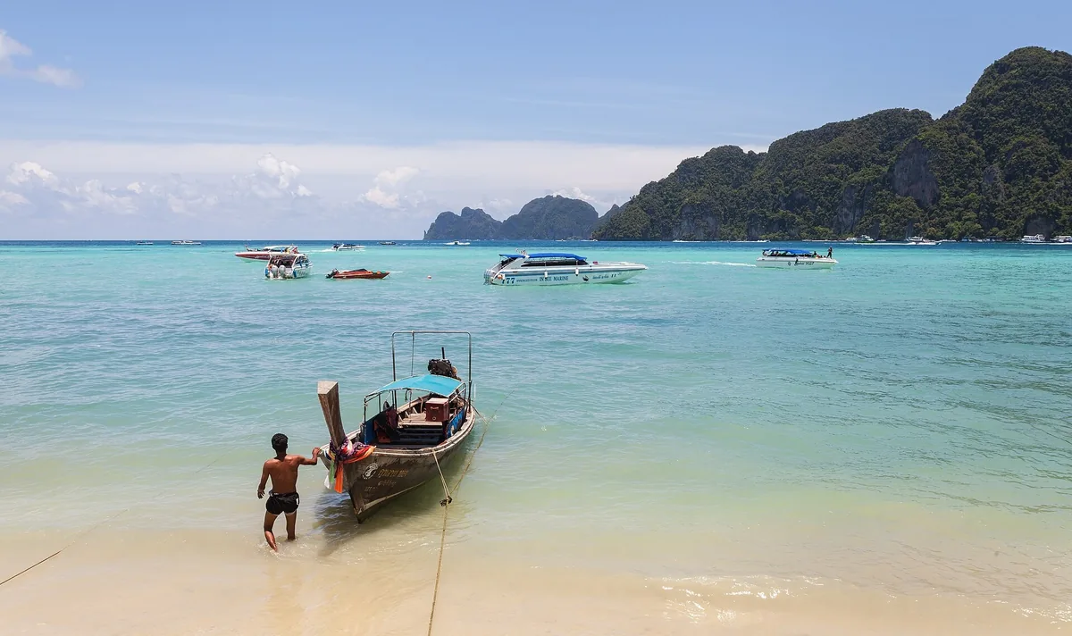 A longtail boat in shallow turquoise water off Ko Phi Phi Don, with speedboats and limestone cliffs behind