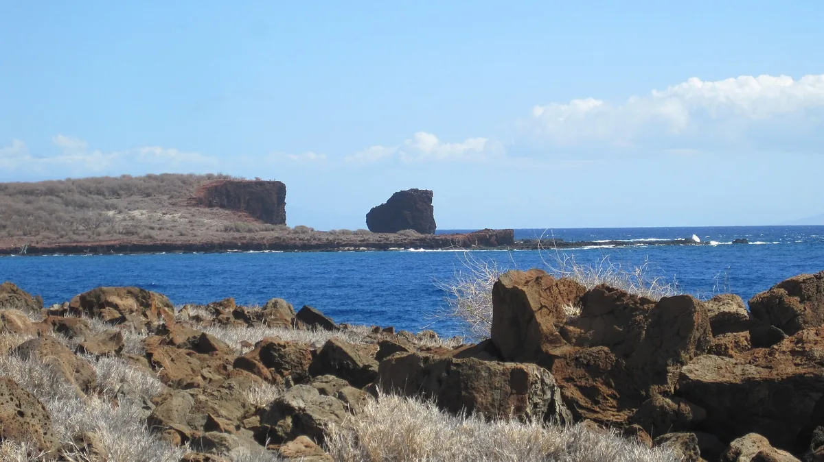 Puʻu Pehe sea stack and the cliffs near Manele seen across deep blue water from the rocky Kapihaʻā shoreline on Lanai