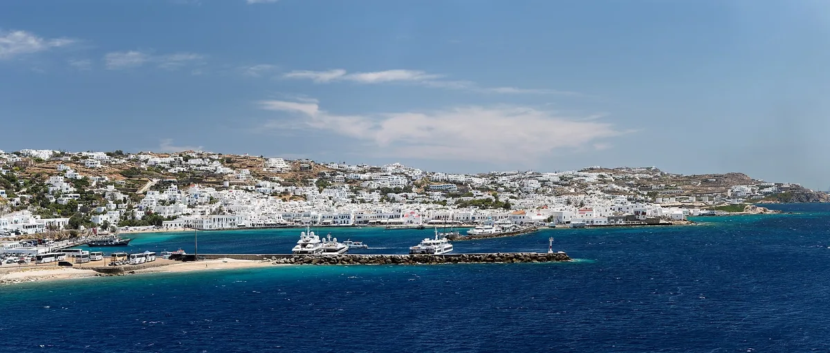 Panorama of the white houses of Mykonos Town spreading along the harbour and hillside, seen across deep blue water