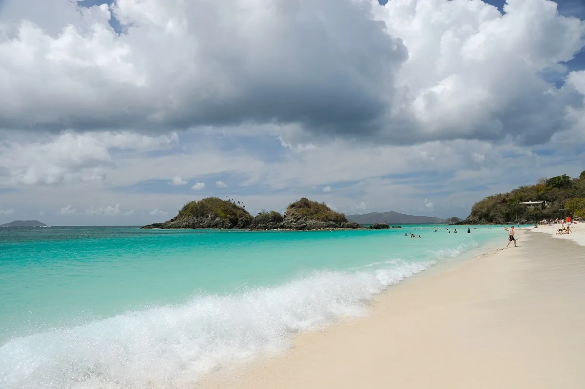 Turquoise surf washing onto the white sand of Trunk Bay on St. John, with Trunk Cay offshore under heavy clouds